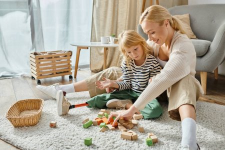 Blondes Mädchen mit Beinprothese sitzt auf Teppich und spielt mit Holzklötzen neben glücklicher Mutter
