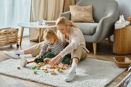 Téléchargez les photos : Fille avec la jambe prothétique assis sur le tapis et jouer jeu de blocs en bois près de mère blonde - en image libre de droit