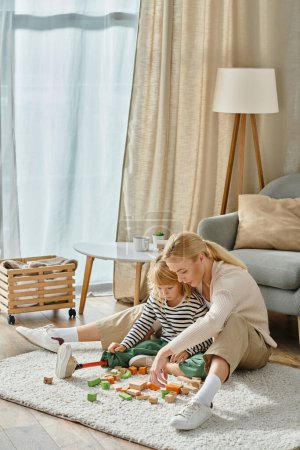 Téléchargez les photos : Fille blonde avec prothèse jambe assise sur le tapis et jouer jeu de blocs en bois près de la mère - en image libre de droit