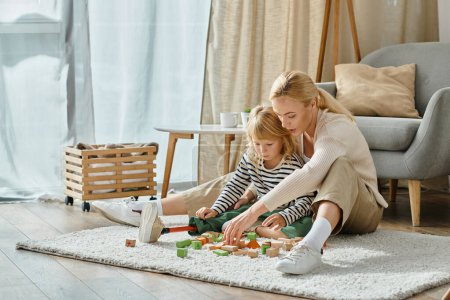 Blondes Mädchen mit Beinprothese sitzt auf Teppich und spielt Holzklötzchen-Spiel in der Nähe fürsorglicher Mutter