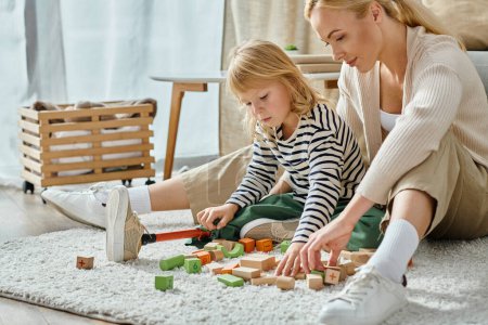 blonde girl with prosthetic leg sitting on carpet and playing with eco wooden blocks near mother