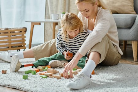 Blondes Mädchen mit Beinprothese sitzt auf Teppich und spielt mit Holzklötzen in der Nähe der hübschen Mutter