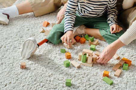 cropped shot, girl with prosthetic leg sitting on carpet and playing with wooden blocks near mother