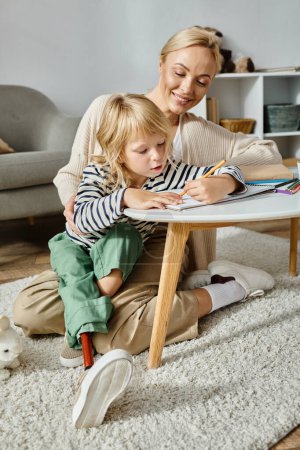 happy blonde woman looking at her daughter with prosthetic leg drawing on paper with colorful pencil