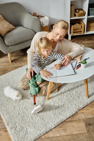 Foto de Mujer rubia feliz mirando a su hija con el dibujo de la pierna protésica en papel con lápiz de colores - Imagen libre de derechos