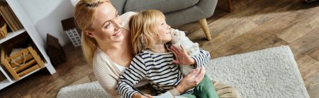 happy woman hugging daughter and looking away while sitting on carpet together, banner