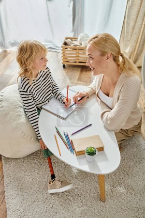 happy mother and kid with prosthetic leg drawing on paper with colorful pencils, quality time