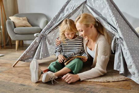 cute girl with prosthetic leg reading book and sitting with happy mother in play tent at home