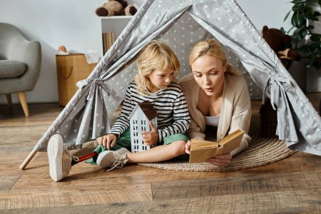 Téléchargez les photos : Fille mignonne avec prothèse jambe et blonde mère lecture livre tout en étant assis dans la tente de jeu à la maison - en image libre de droit