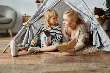 Téléchargez les photos : Petite fille avec prothèse jambe et blonde mère lecture livre et assis dans la tente de jeu à la maison - en image libre de droit
