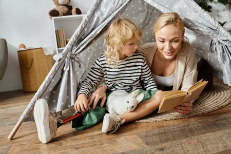 Téléchargez les photos : Petite fille avec prothèse jambe tenant jouet doux près de mère lecture livre dans la tente de jeu à la maison - en image libre de droit