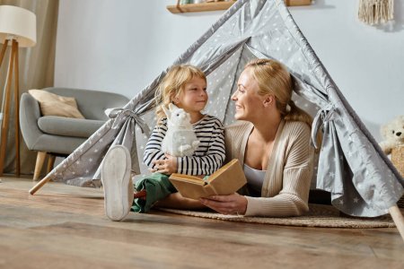 Téléchargez les photos : Fille souriante avec prothèse jambe tenant jouet doux près de la mère livre de lecture dans la tente de jeu à la maison - en image libre de droit