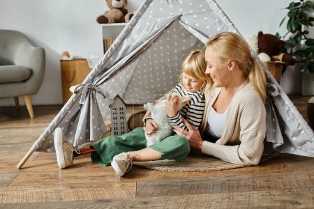 Téléchargez les photos : Petite fille avec prothèse jambe étreignant lapin jouet doux près de mère heureuse tout en étant assis dans la tente de jeu - en image libre de droit