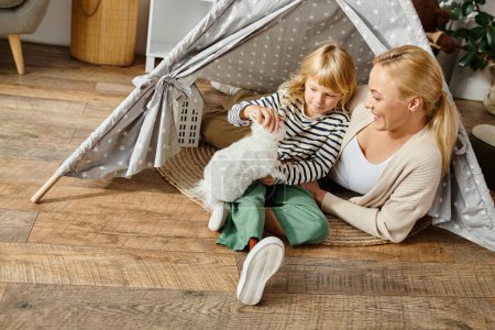 Téléchargez les photos : Fille heureuse avec prothèse jambe jouer avec doux jouet lapin près de mère tout en étant assis dans la tente de jeu - en image libre de droit