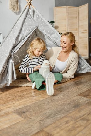 Téléchargez les photos : Fille blonde avec prothèse jambe jouer avec doux jouet lapin près de mère tout en étant assis dans la tente de jeu - en image libre de droit