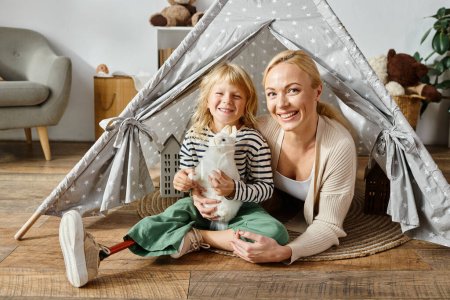 Téléchargez les photos : Portrait de famille de fille heureuse avec la jambe prothétique tenant jouet doux près de la mère dans la tente de jeu - en image libre de droit