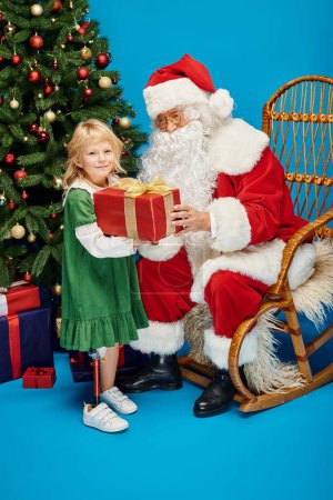 Foto de Santa dando regalo a la chica feliz con la pierna protésica al lado del árbol de Navidad en el fondo azul - Imagen libre de derechos