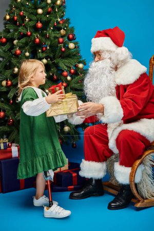 Foto de Santa Claus dando regalos a chica feliz con la pierna protésica al lado del árbol de Navidad en azul - Imagen libre de derechos