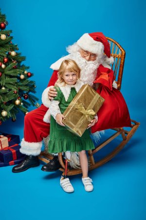 Foto de Santa Claus dando regalo a la chica feliz con la pierna protésica al lado del árbol de Navidad en azul - Imagen libre de derechos