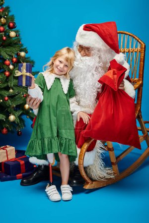 Foto de Alegre chica con prótesis pierna celebración presente cerca de Santa Claus junto a árbol de Navidad en azul - Imagen libre de derechos