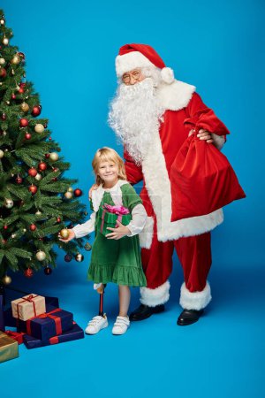 Foto de Santa Claus bolsa de mano cerca de chica alegre con la pierna protésica al lado del árbol de Navidad en azul - Imagen libre de derechos