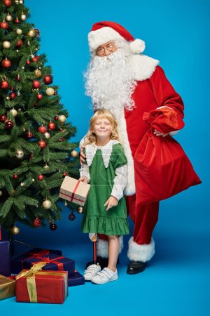 Foto de Santa Claus sosteniendo bolsa de saco cerca de chica feliz con la pierna protésica junto al árbol de Navidad en azul - Imagen libre de derechos