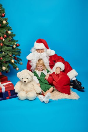 Foto de Chica feliz con la pierna protésica y el oso de peluche y sentado con Santa Claus al lado del árbol de Navidad - Imagen libre de derechos