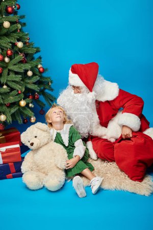 happy kid with prosthetic leg and teddy bear sitting with Santa Claus next to Christmas tree