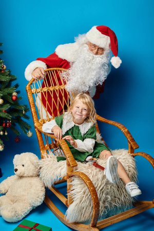 Foto de Santa Claus mecedora con chica feliz con pierna protésica al lado de oso de peluche y árbol de Navidad - Imagen libre de derechos