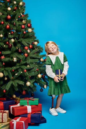 cheerful little girl with prosthetic leg holding wrapped present next to Christmas tree on blue