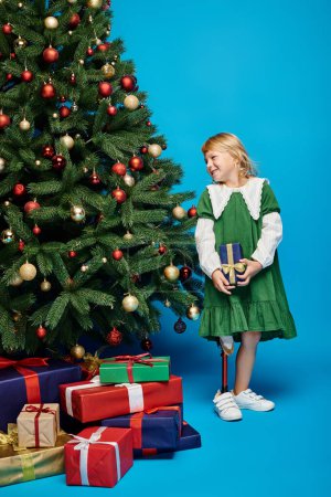 joyful little girl with prosthetic leg holding wrapped present next to Christmas tree on blue