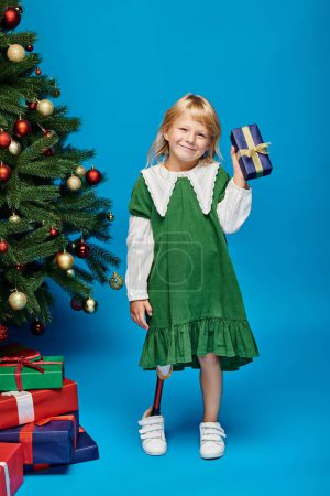 pleased little girl with prosthetic leg holding wrapped present next to Christmas tree on blue