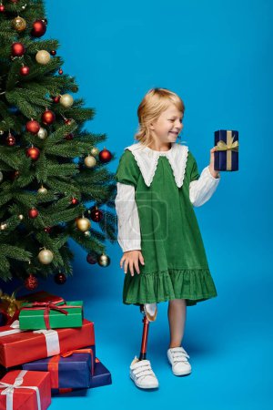 positive little girl with prosthetic leg holding wrapped present next to Christmas tree on blue
