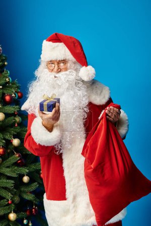 Santa Claus con barba y anteojos en traje rojo sosteniendo saco y regalo cerca del árbol de Navidad