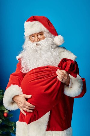 Père Noël avec barbe et lunettes tenant sac rouge avec cadeaux de Noël sur bleu