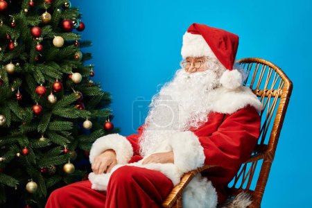 happy Santa Claus with beard and eyeglasses sitting in rocking chair near Christmas tree on blue