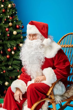 cheerful Santa Claus with beard and eyeglasses sitting in rocking chair near Christmas tree on blue