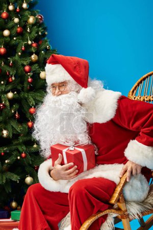 heureux Père Noël avec barbe et lunettes assis dans un fauteuil à bascule avec cadeau près de l'arbre de Noël