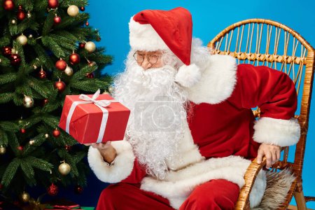 happy Santa Claus with beard and eyeglasses sitting in rocking chair with gift near Christmas tree