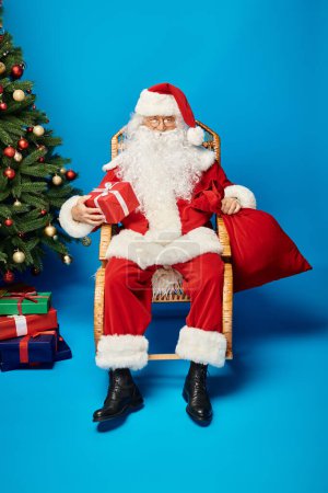 happy Santa Claus in eyeglasses sitting in rocking chair with gift and sack bag near Christmas tree