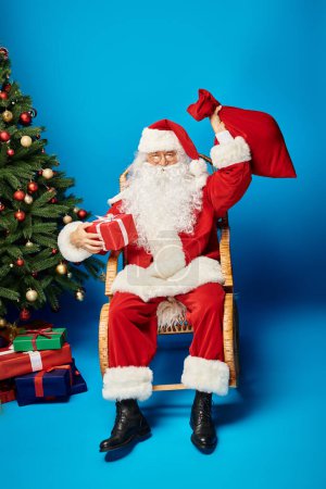 happy Santa Claus in eyeglasses sitting in rocking chair with gift and sack bag near Christmas tree