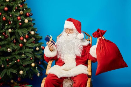 happy Santa Claus sitting in rocking chair with gift box and sack bag near Christmas tree on blue