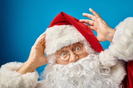 Porträt eines aufgeregten Weihnachtsmannes mit weißem Bart und Brille, der auf blauem Hintergrund in die Kamera blickt