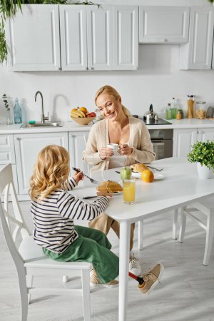 Téléchargez les photos : Mère blonde souriante regardant sa fille mignonne avec une jambe prothétique petit déjeuner dans la cuisine - en image libre de droit