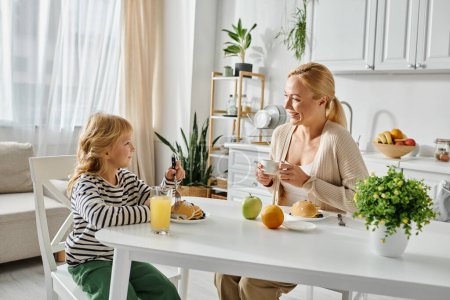 Téléchargez les photos : Joyeuse mère blonde regardant sa fille mignonne avec jambe prothétique prendre le petit déjeuner dans la cuisine - en image libre de droit