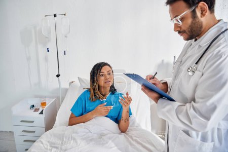 Téléchargez les photos : Jeune médecin indien regardant un patient afro-américain couché dans un service hospitalier, soins de santé - en image libre de droit