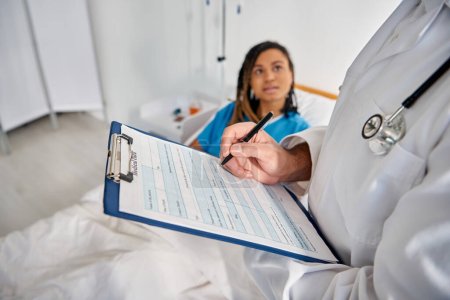 Téléchargez les photos : Attrayant afro-américain femme couchée dans le lit dans la salle d'hôpital parler à son jeune médecin indien - en image libre de droit