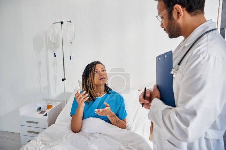 Téléchargez les photos : Attrayant afro-américain femme couchée dans le lit dans la salle d'hôpital parler à son jeune médecin indien - en image libre de droit