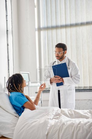 Téléchargez les photos : Beau médecin indien en lunettes parlant à un jeune patient afro-américain à l'hôpital, soins de santé - en image libre de droit