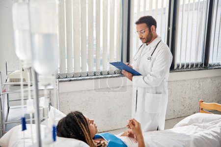 Téléchargez les photos : Beau médecin indien prenant des notes sur le patient afro-américain couché dans le lit dans la salle d'hôpital - en image libre de droit